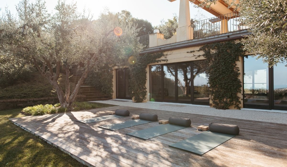 Outdoor yoga setup with mats, bolsters, and blocks on a wooden deck beside a house covered in ivy, under sunlight and olive trees.