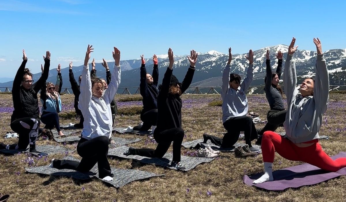 Group of people practicing yoga on mats outdoors with raised arms, set against a backdrop of snow-capped mountains and clear blue sky.