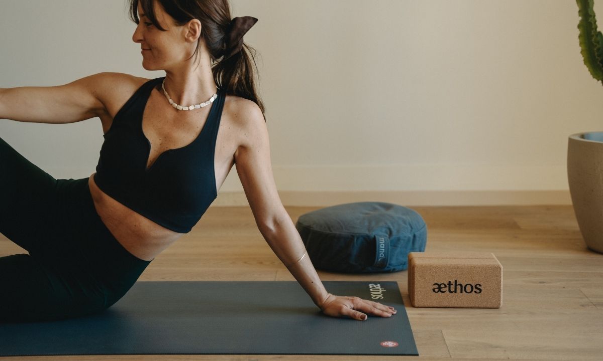 Woman in black workout clothes doing yoga on a mat next to a cushion and a cork yoga block in a minimalist room.