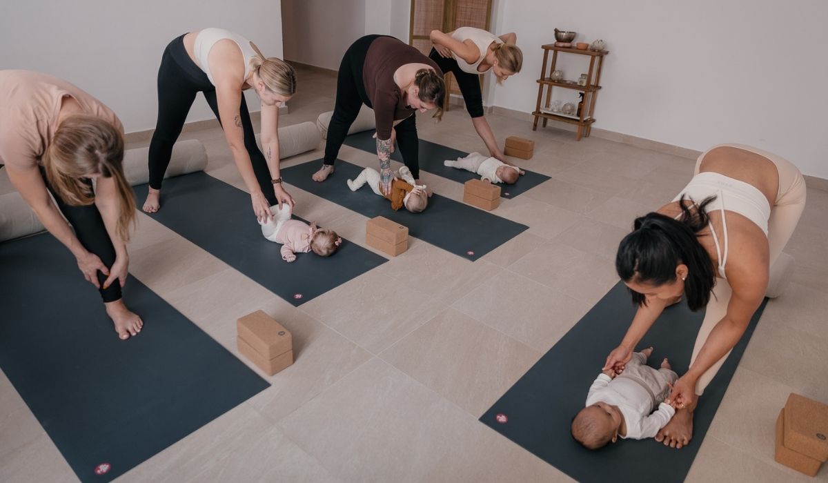 Mothers practicing yoga stretches on mats while interacting with their babies lying in front of them in a calm studio setting.