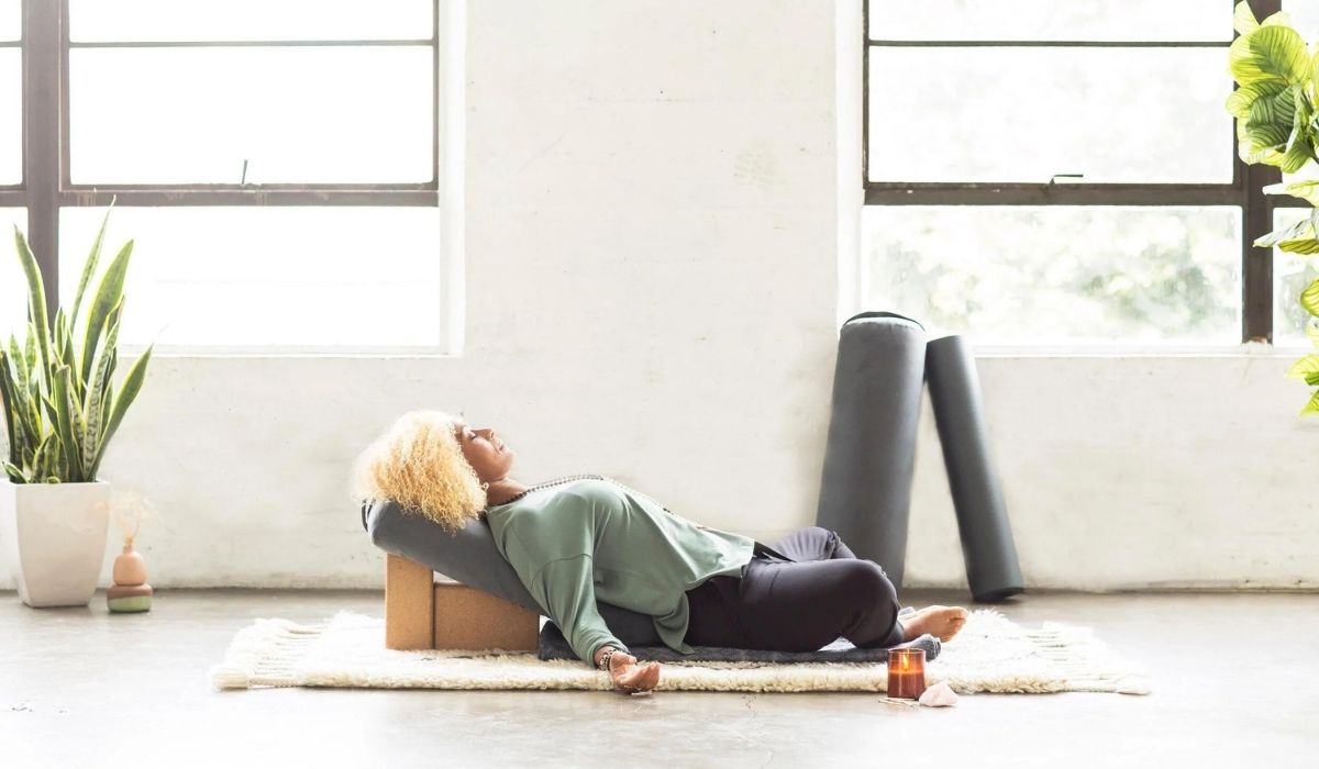A woman with curly hair relaxes on a yoga mat with props, surrounded by plants and natural light from large windows.