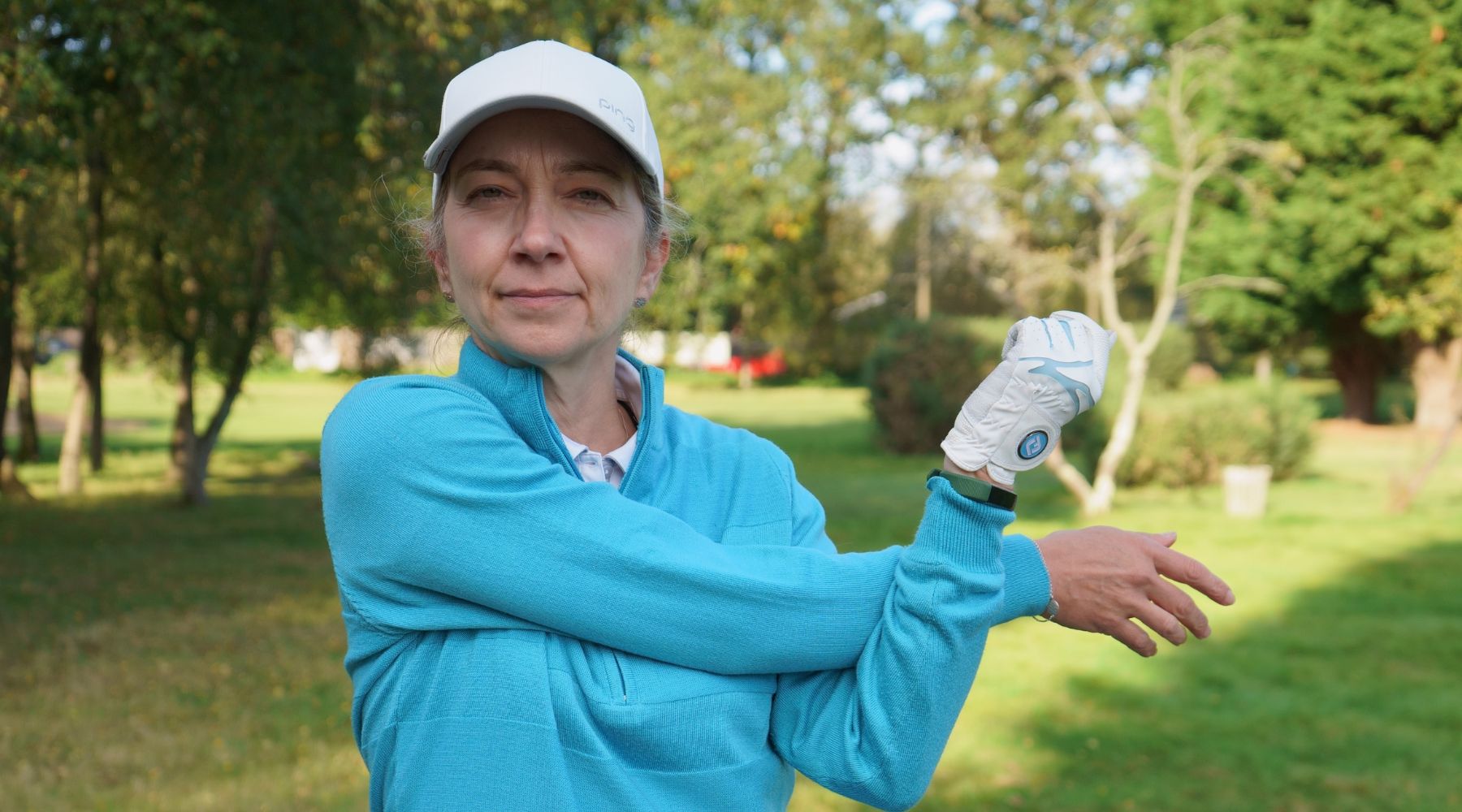 A person in a blue sweater and golf gloves stretches their arm in a grassy park, surrounded by trees.