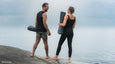 A man and woman stand barefoot on a rocky shore, holding Manduka yoga mats, gazing at a calm ocean under a cloudy sky.