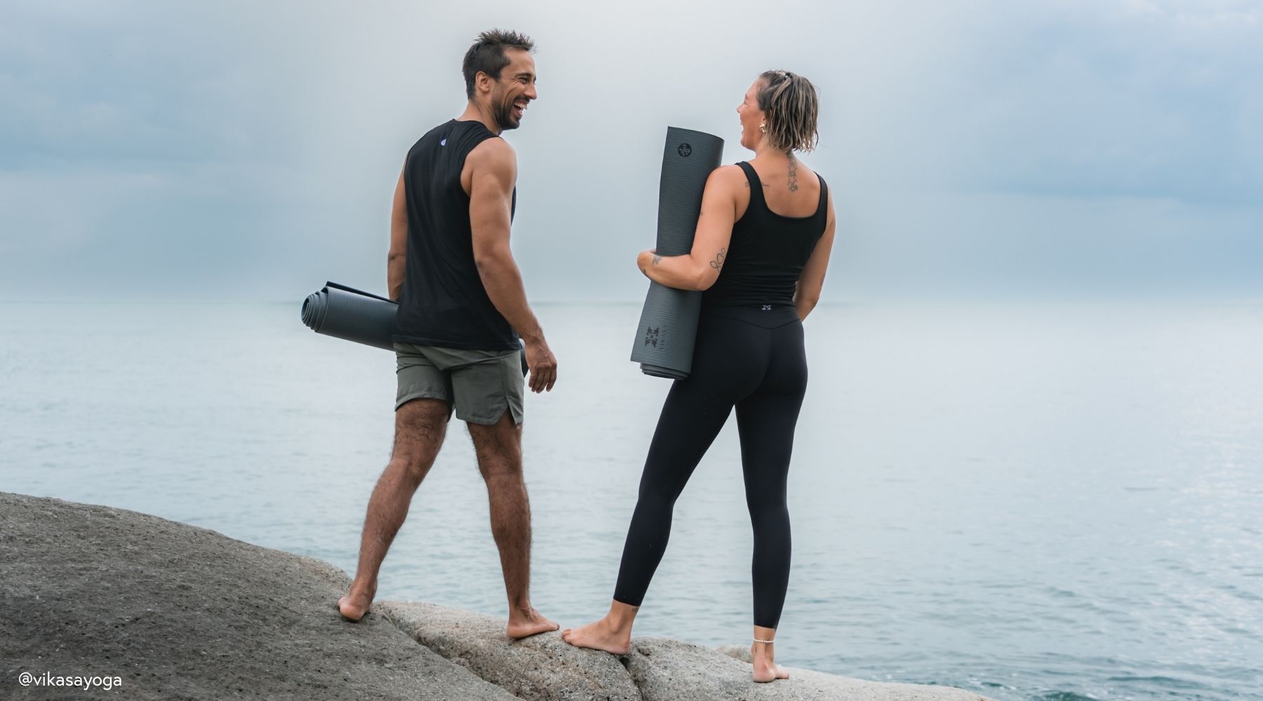 A man and woman stand barefoot on a rocky shore, holding Manduka yoga mats, gazing at a calm ocean under a cloudy sky.