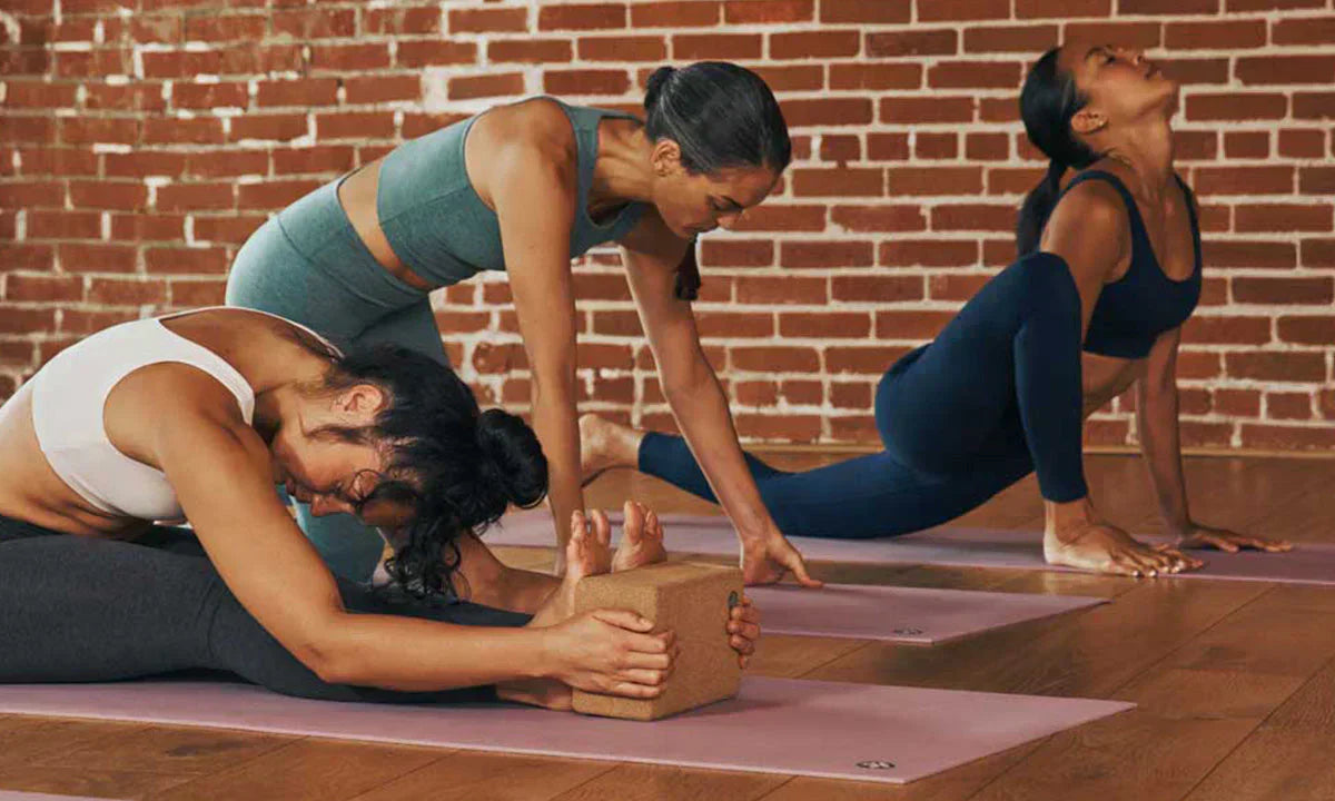 Three women practice yoga poses on mats in a warm studio with a brick wall, focusing on flexibility and balance.