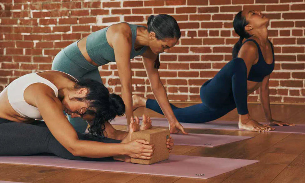 Three women practice yoga poses on mats in a warm studio with a brick wall, focusing on flexibility and balance.