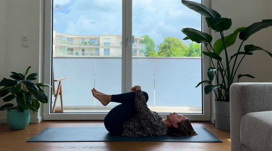 A person in comfortable clothing lies on a yoga mat, practicing a pose near a large balcony window with plants in the foreground.
