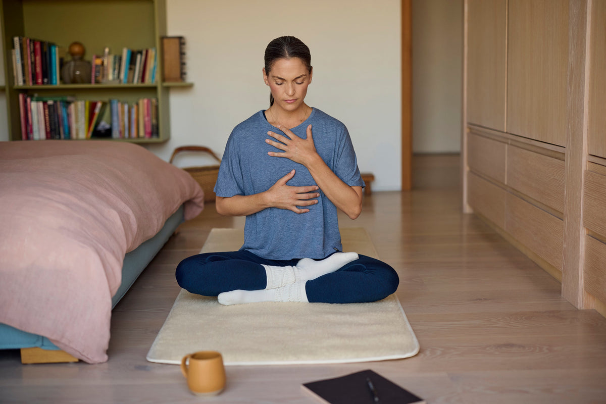 A person sits cross-legged on a mat, practicing mindfulness or meditation in a cozy, well-lit room with a bed and bookshelves.