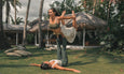 Two women practice an acrobatic yoga pose outdoors, surrounded by palm trees and a thatched-roof structure, on a grassy area.
