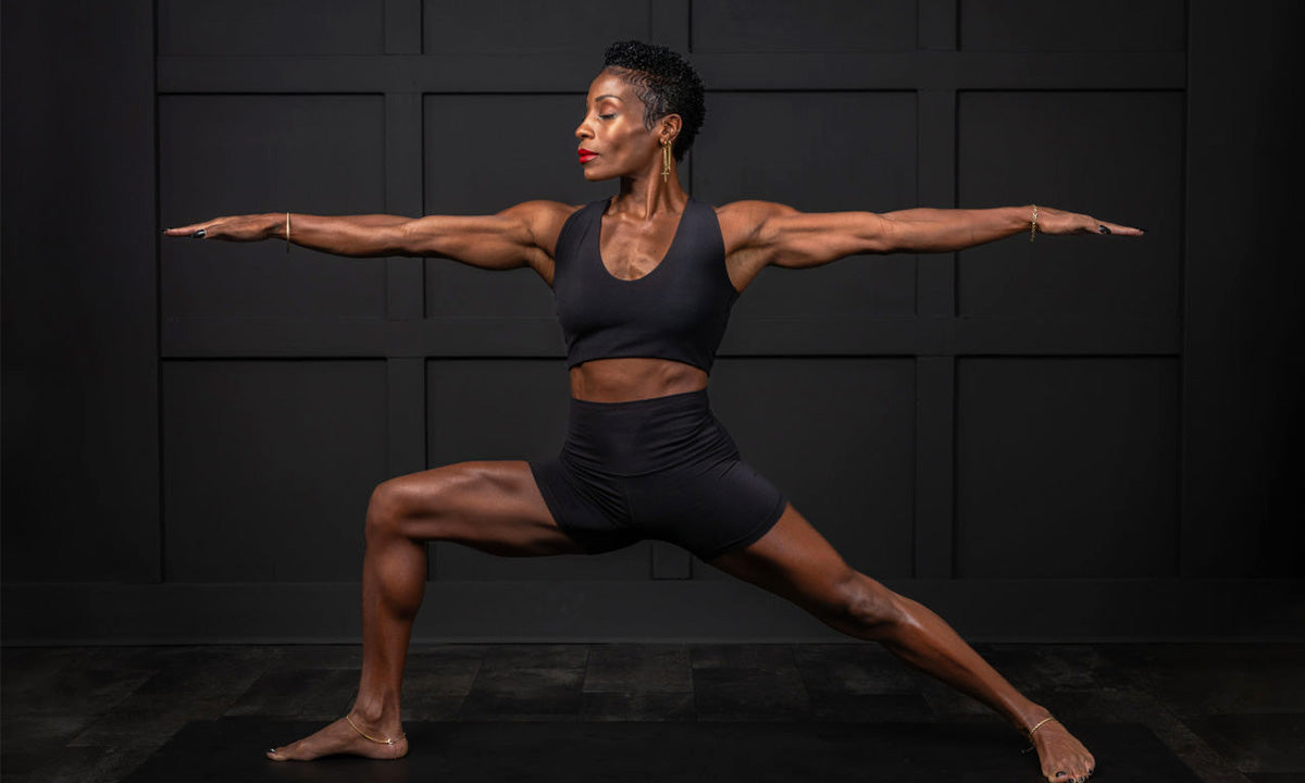 A woman is demonstrating a warrior yoga pose on a black mat against a dark wall, showcasing strength and balance.