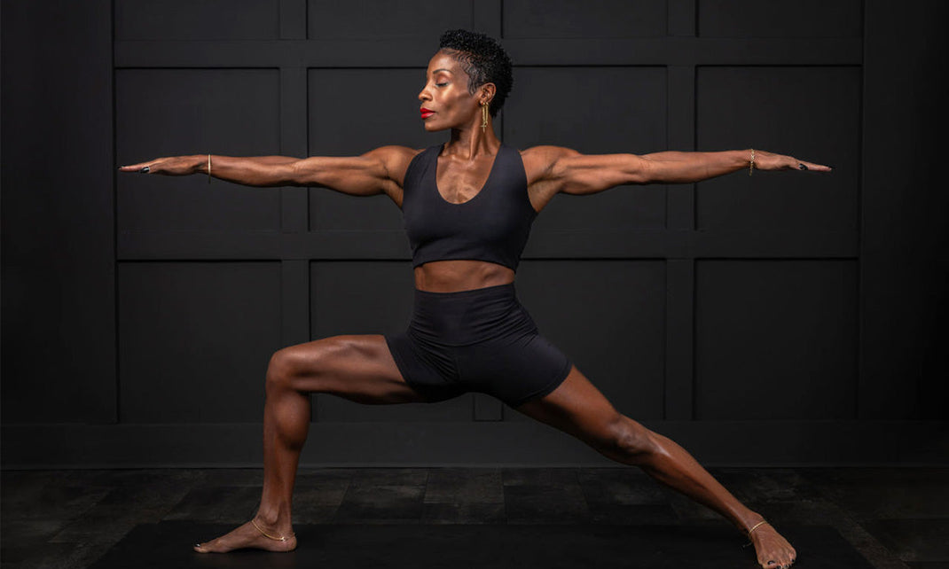 A woman is demonstrating a warrior yoga pose on a black mat against a dark wall, showcasing strength and balance.