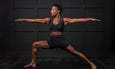 A woman is demonstrating a warrior yoga pose on a black mat against a dark wall, showcasing strength and balance.