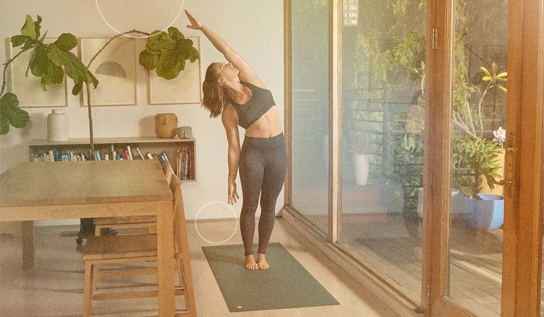 A woman in athletic wear stretches on a yoga mat inside a bright, airy room with a wooden table and plants in the background.