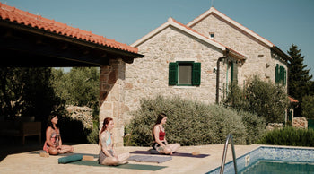 Three women practice yoga on colorful mats by a stone house, surrounded by olive trees and a serene swimming pool under a clear blue sky.