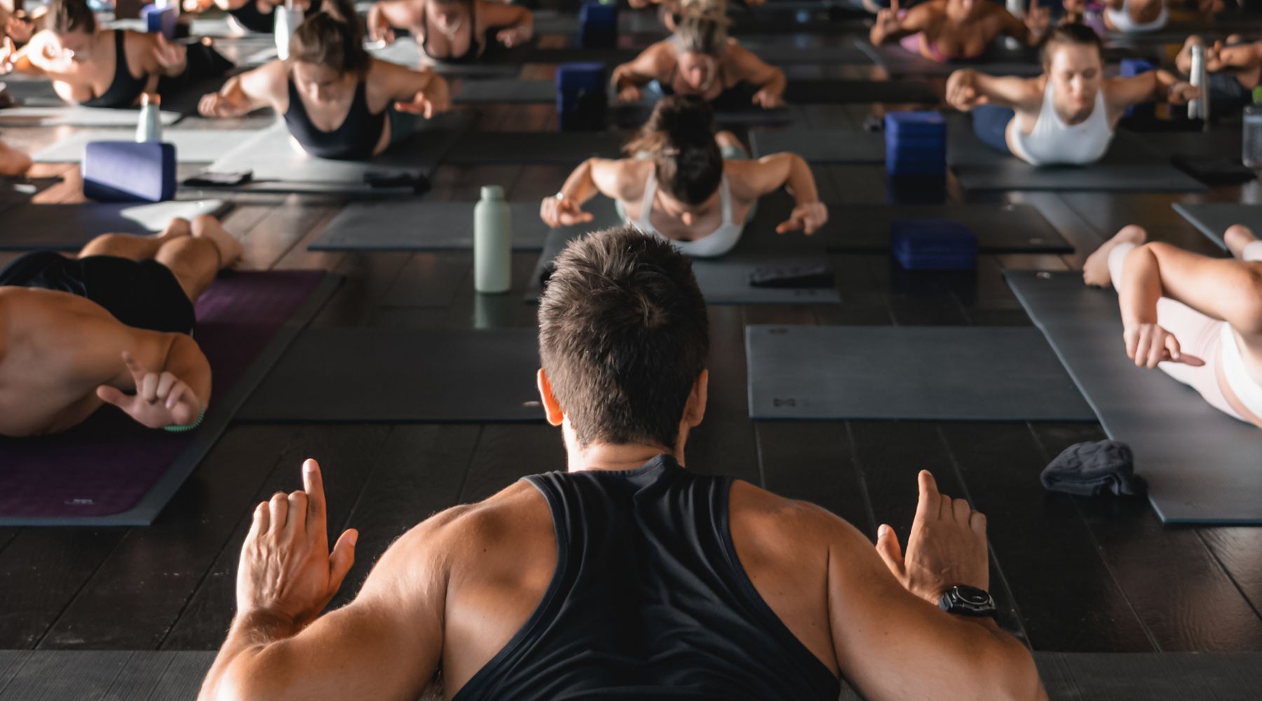 A yoga instructor leads a group yoga class, with participants in various poses on mats in a spacious, well-lit studio.