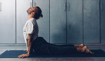 A person performs the Cobra pose on a yoga mat in a minimalist indoor space with gray walls and cabinets.