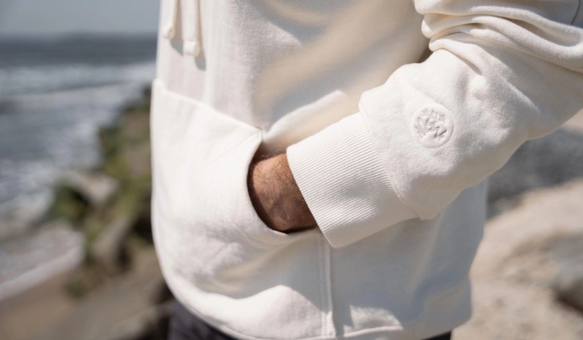 A person wearing a cream-colored sweatshirt with a logo on the sleeve, standing near the beach with ocean waves in the background.