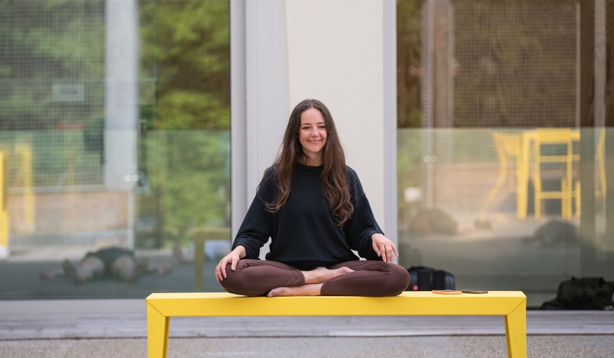 A person sits cross-legged on a bright yellow bench, surrounded by greenery and glass reflections, exuding a serene atmosphere.