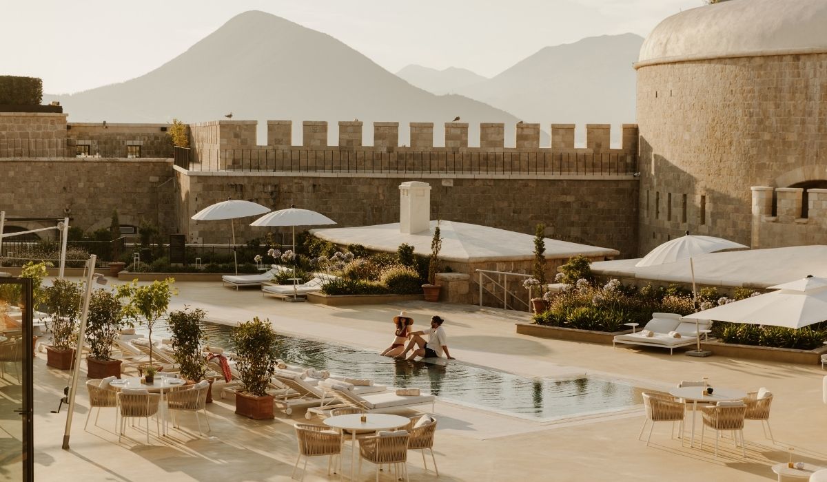 Outdoor pool area with stone castle walls, mountains in background, lounge chairs, umbrellas, and a couple sitting by the water.