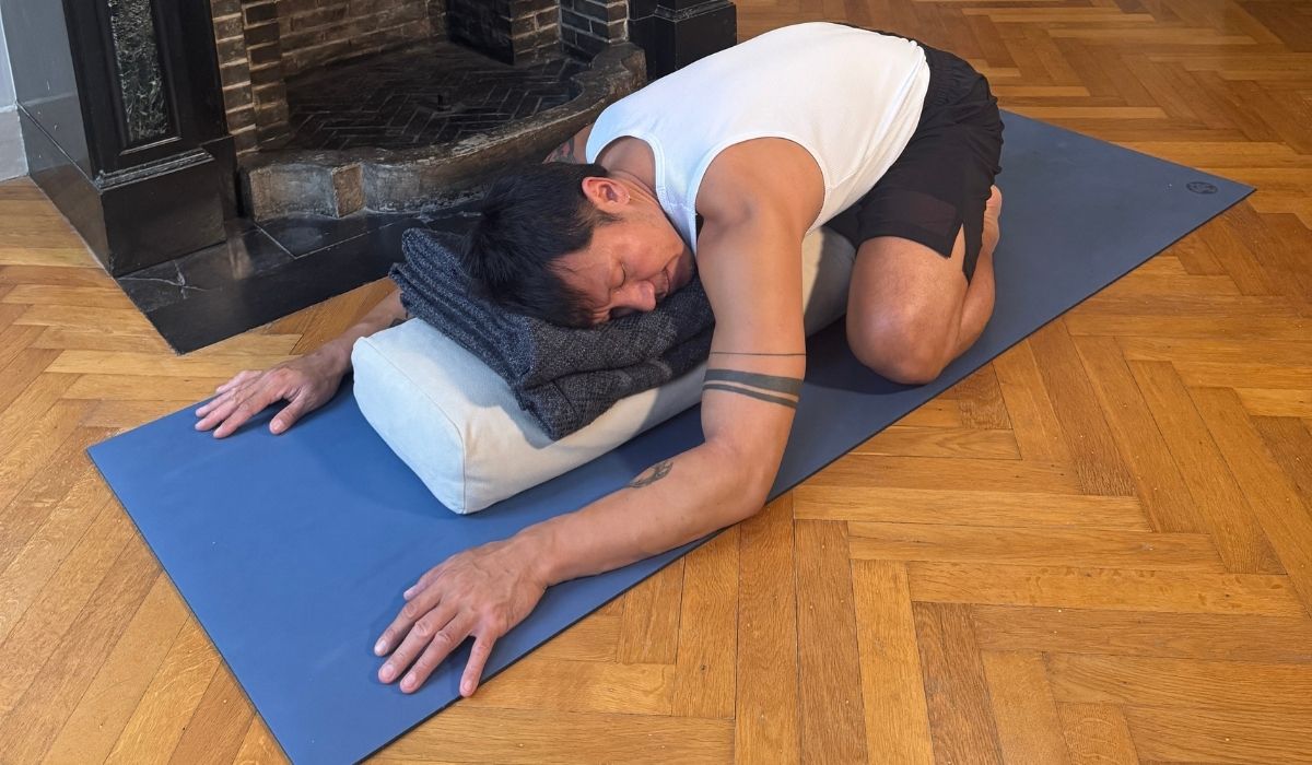 A person in a white tank top and shorts performs a yoga pose on a blue mat, resting on cushions in a cozy room.