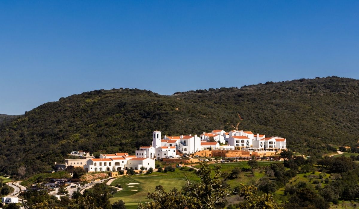 A picturesque white-walled resort nestled on a hillside, surrounded by green vegetation under a clear blue sky.