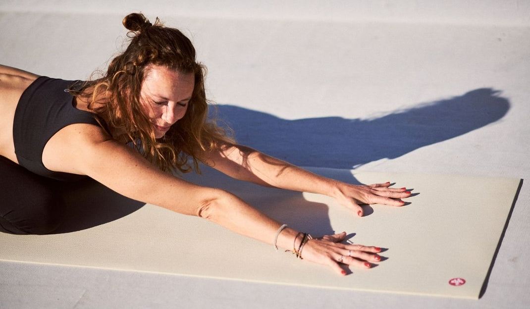 A person practices yoga on a manduka yoga mat outdoors, stretching forward with arms extended, surrounded by natural light and soft shadows.