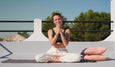 A person sits cross-legged on a rooftop, practicing yoga with hands in a prayer position, surrounded by soft pillows and greenery.