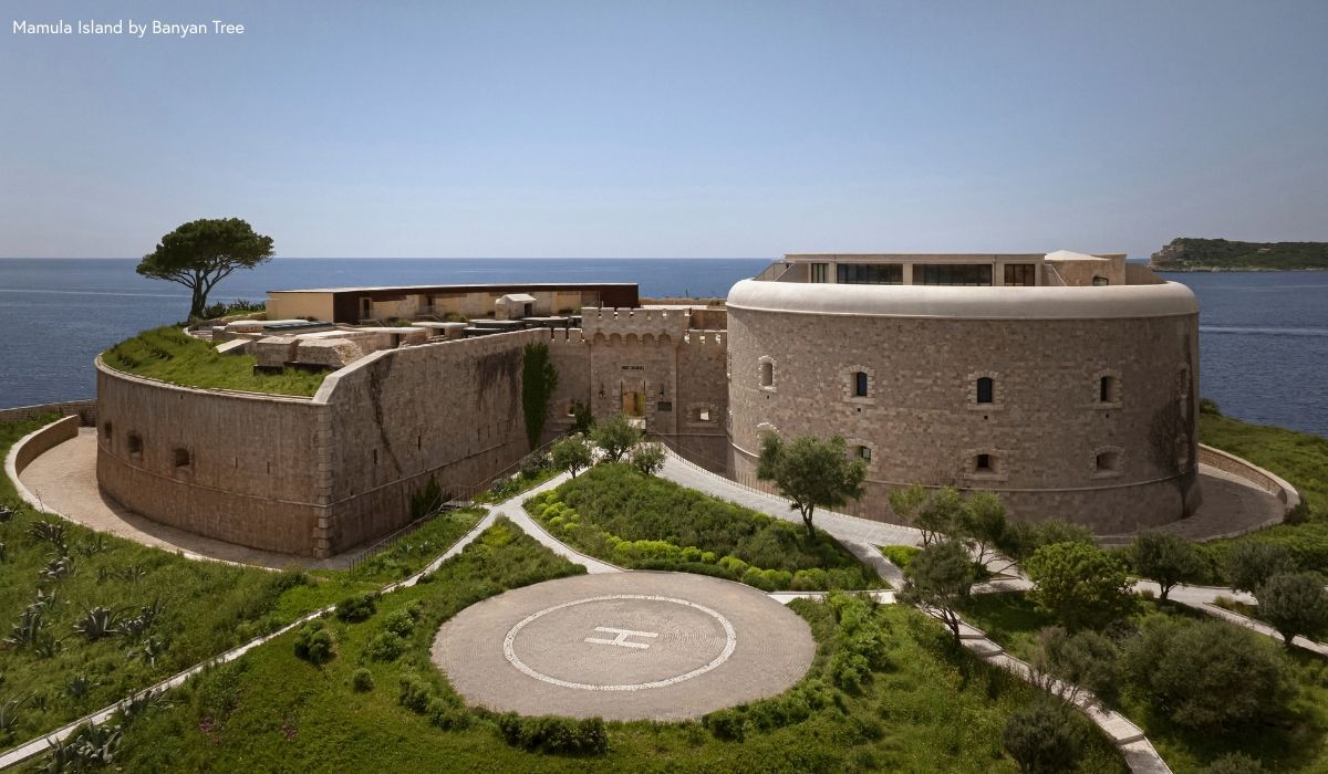 Historic stone fort with rounded tower by the sea, surrounded by greenery and a helipad in the foreground under a clear blue sky.