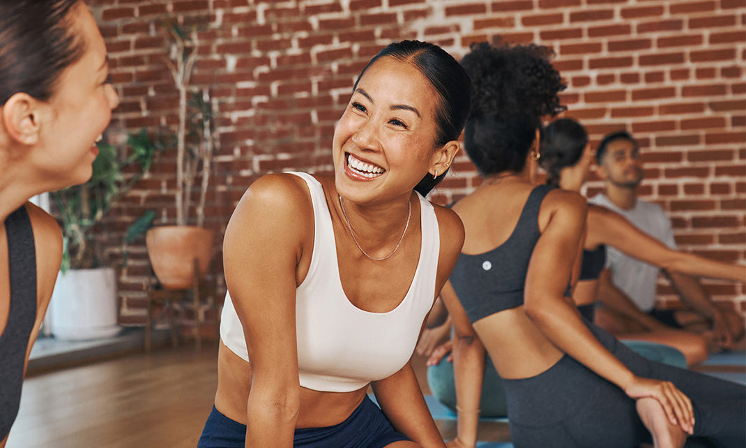 A group of four people in manduka athletic wear seated in a yoga studio with a brick wall, engaged in a meditative or reflective moment.