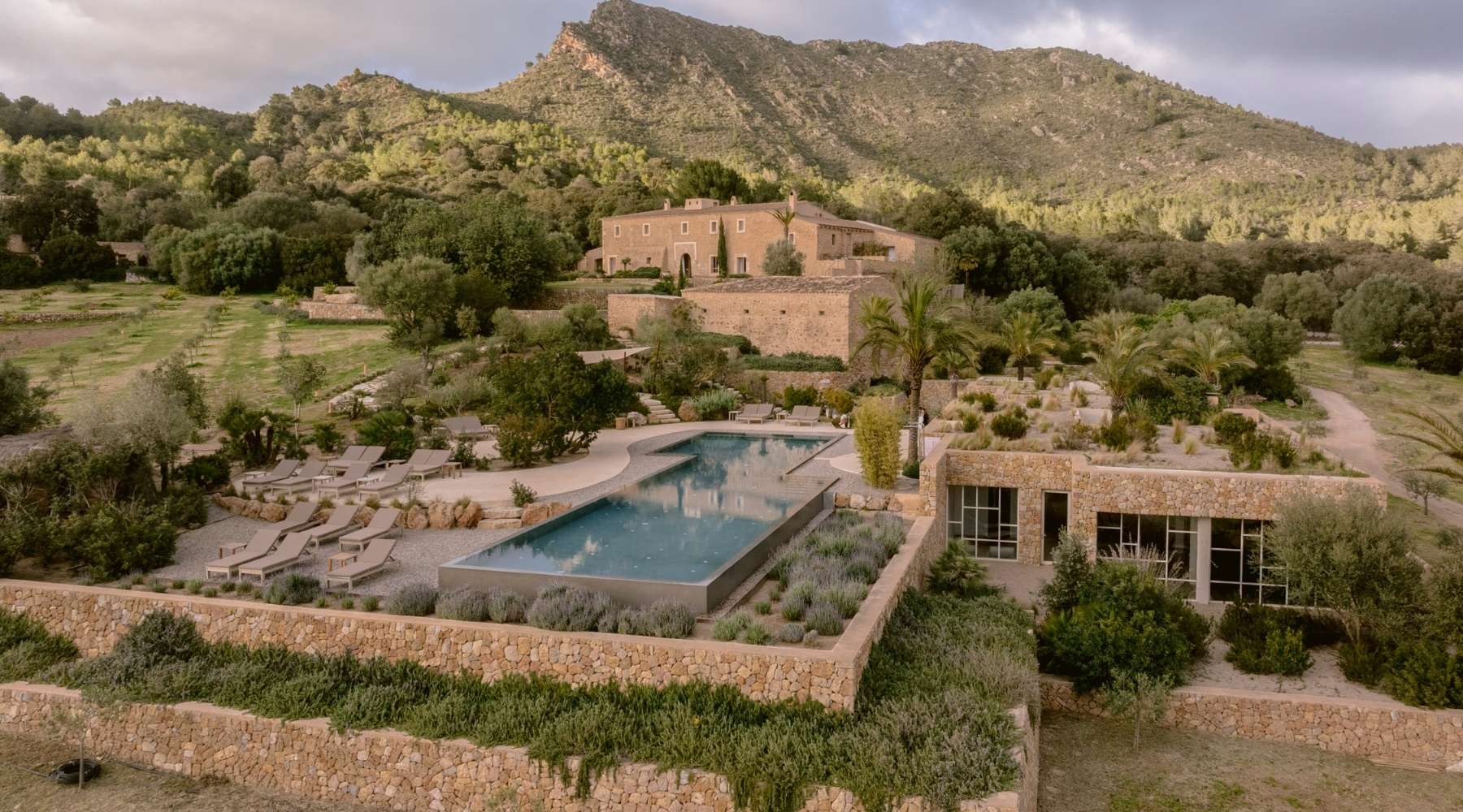 A serene landscape featuring a luxury villa, tranquil pool, and sun loungers, framed by lush greenery and mountains under a cloudy sky.