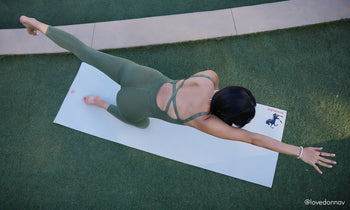 A person performs a yoga pose on a mat in an outdoor setting, wearing a fitted olive green outfit, with one arm extended forward.