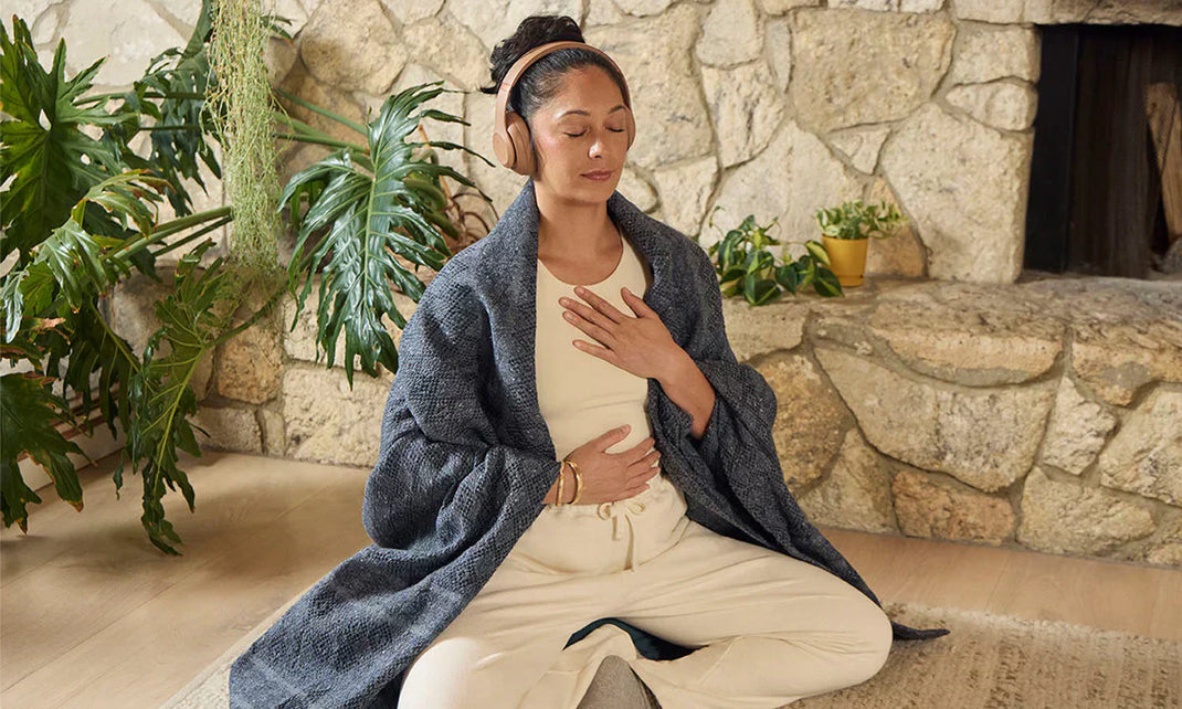 A person sits cross-legged on the floor, hands on their stomach, wearing a cozy blanket and headphones, surrounded by indoor plants.
