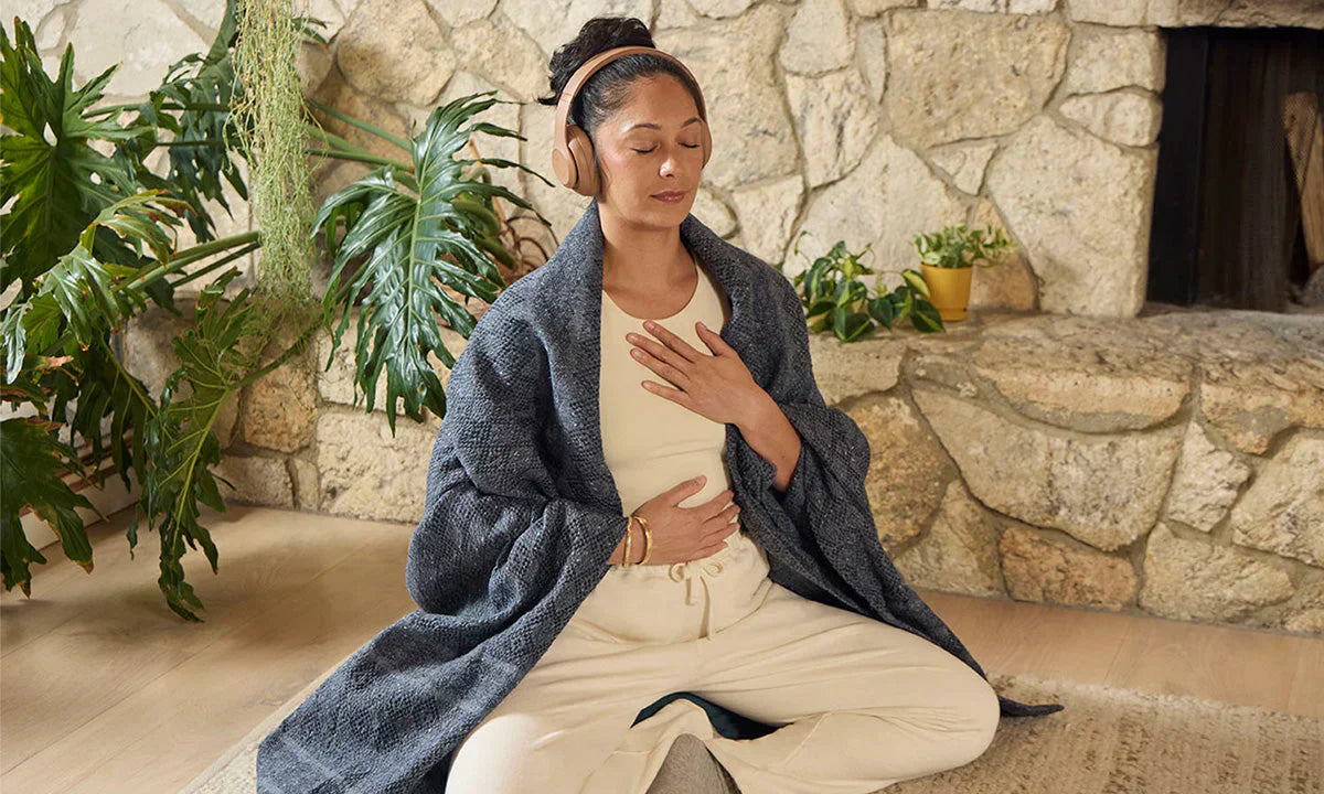 A person sits cross-legged on the floor, hands on their stomach, wearing a cozy blanket and headphones, surrounded by indoor plants.