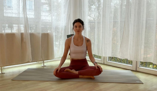 A person sits cross-legged on a yoga mat in a sunlit room, surrounded by sheer curtains and large windows letting in natural light.