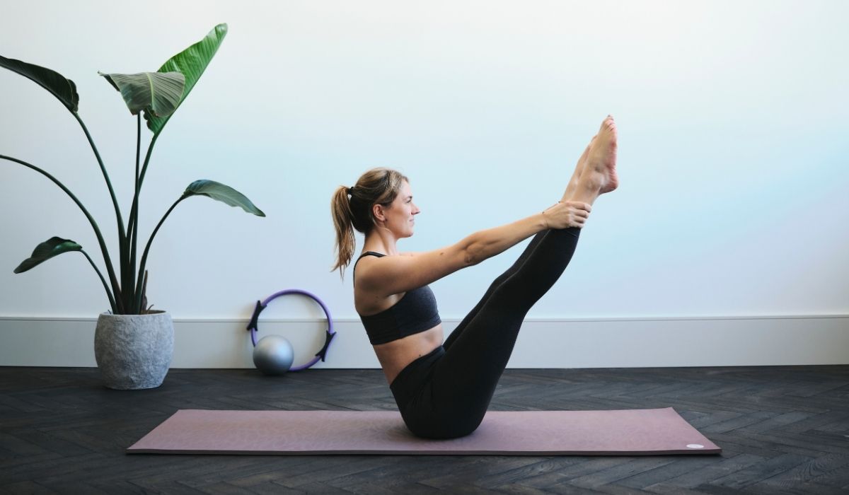 Woman in black workout clothes practicing yoga, balancing on a mat in a bright room with a potted plant and exercise equipment.
