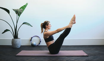 Woman in black workout clothes practicing yoga, balancing on a mat in a bright room with a potted plant and exercise equipment.