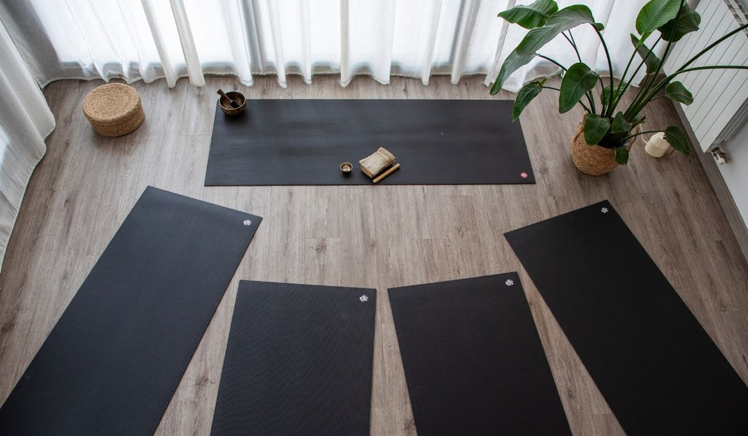 Five black yoga mats arranged on a wood floor in a bright room with sheer curtains, a potted plant, and meditation accessories.