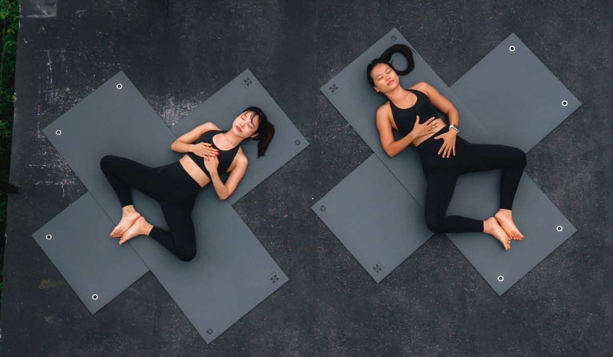 Two women in black workout clothes relax on gray yoga mats arranged in an "X" shape on a dark surface.