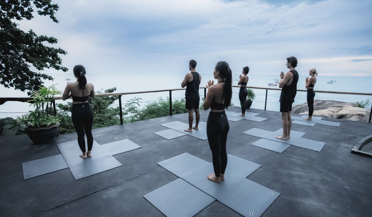 Group of six people practicing yoga on mats on a terrace overlooking the sea during early morning or dusk.