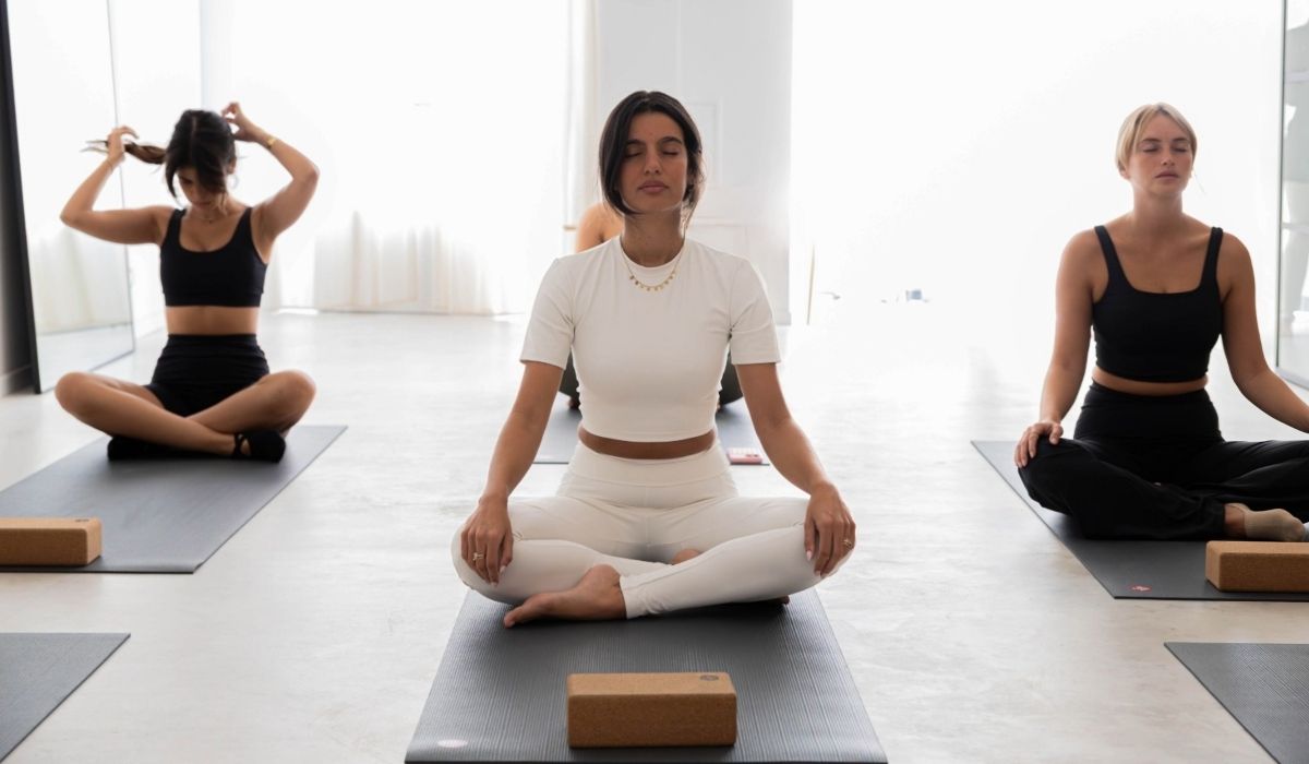 Three women sit cross-legged on yoga mats in a bright studio, meditating and preparing for a yoga session.