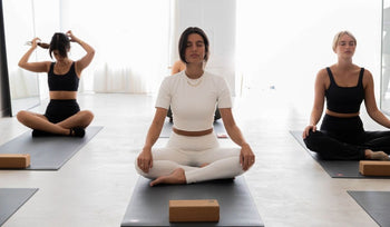 Three women sit cross-legged on yoga mats in a bright studio, meditating and preparing for a yoga session.