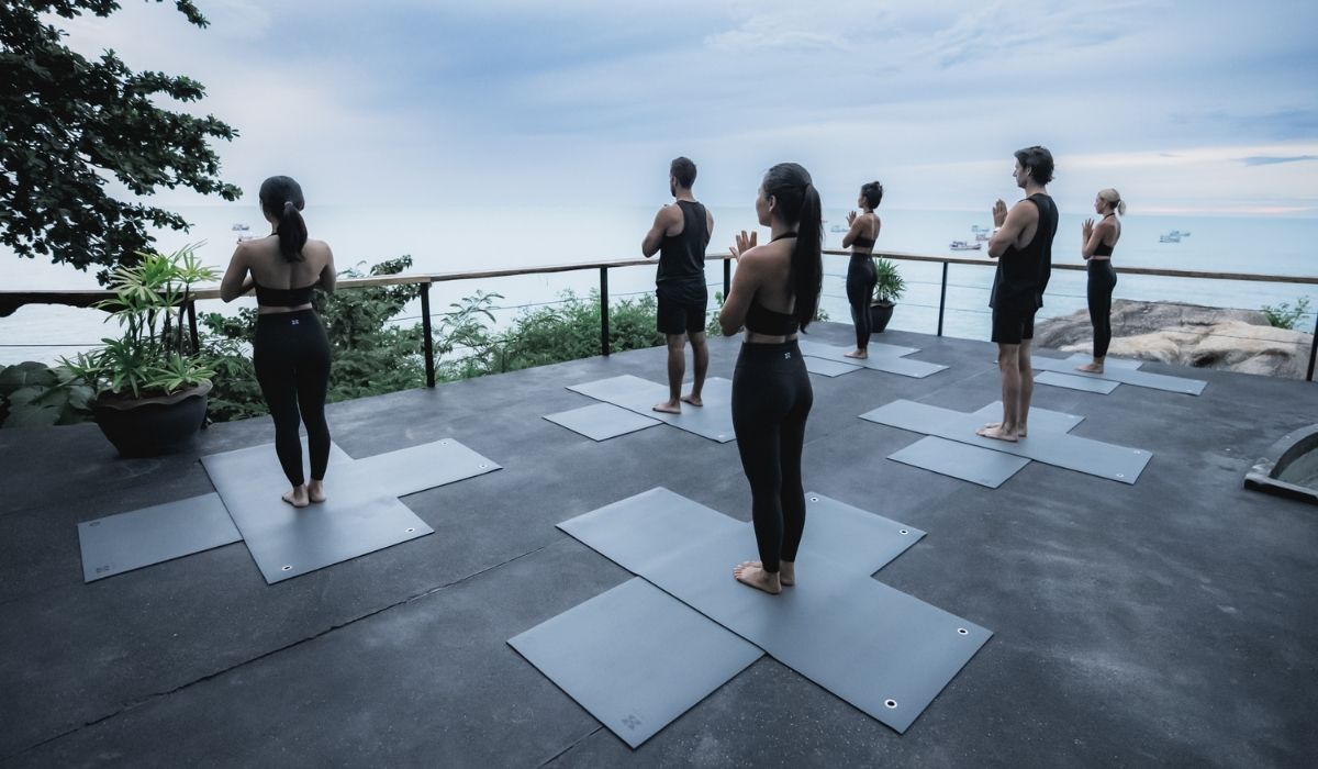Group of people practicing yoga with hands in prayer pose on mats overlooking the ocean at sunrise.