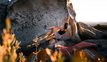 Woman bouldering on a large rock outdoors with crash pads below, during sunset or sunrise.