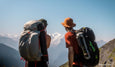 Two hikers with large backpacks overlooking distant snow-capped mountains under a clear blue sky.