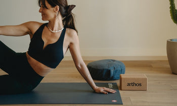 Woman in black workout clothes doing yoga on a mat next to a cushion and a cork yoga block in a minimalist room.