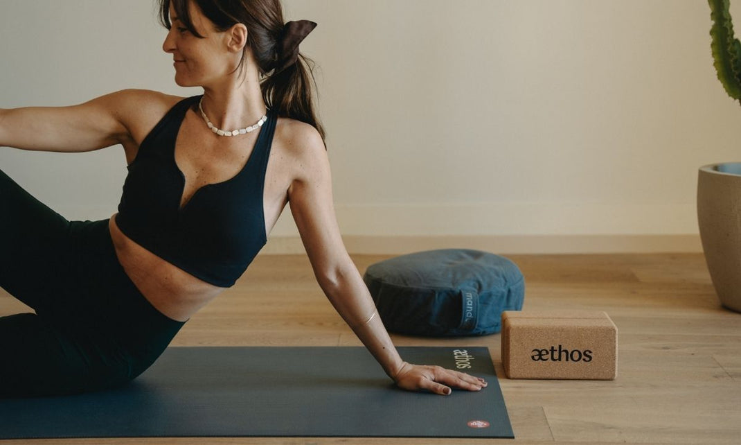Woman in black workout clothes doing yoga on a mat next to a cushion and a cork yoga block in a minimalist room.