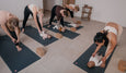 Mothers practicing yoga stretches on mats while interacting with their babies lying in front of them in a calm studio setting.