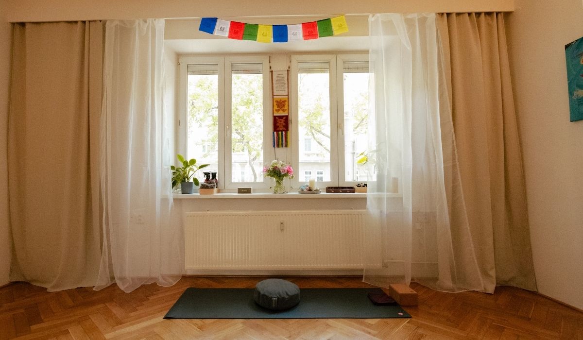 Calm yoga space with mat, meditation cushion, blocks, sheer curtains, potted plants, flowers, and colorful prayer flags by a large window.