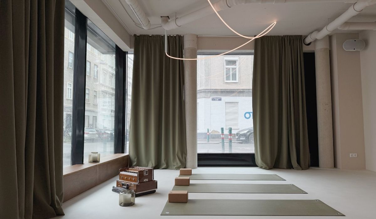 Minimalist yoga studio with green mats, cork blocks, and a harmonium near large windows with green curtains.