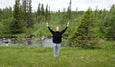 Person standing on grass with arms raised near a flowing river surrounded by green trees and forest under a cloudy sky.